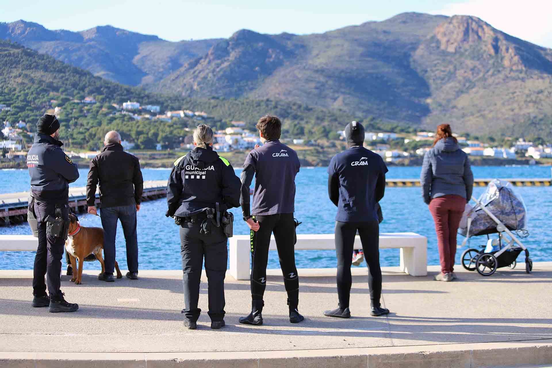 Una miembro de la Guardia Municipal, otro de la Unidad Marítima de los Mossos de Escuadra, técnicos del CRAM y varias personas observan detenidamente el ejemplar de delfín listado en el Moll d’en Balleu en el Port de la Selva en Catalunya (España) / Foto: FFM - EA