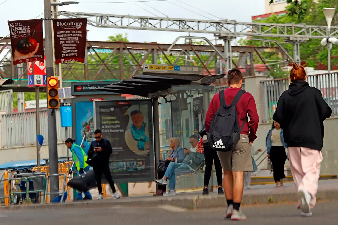 Personas esperan en una parada de autobús en La Farga de L’Hospitalet bajo un cartel de BioCultura Barcelona 2026 / Foto: EA