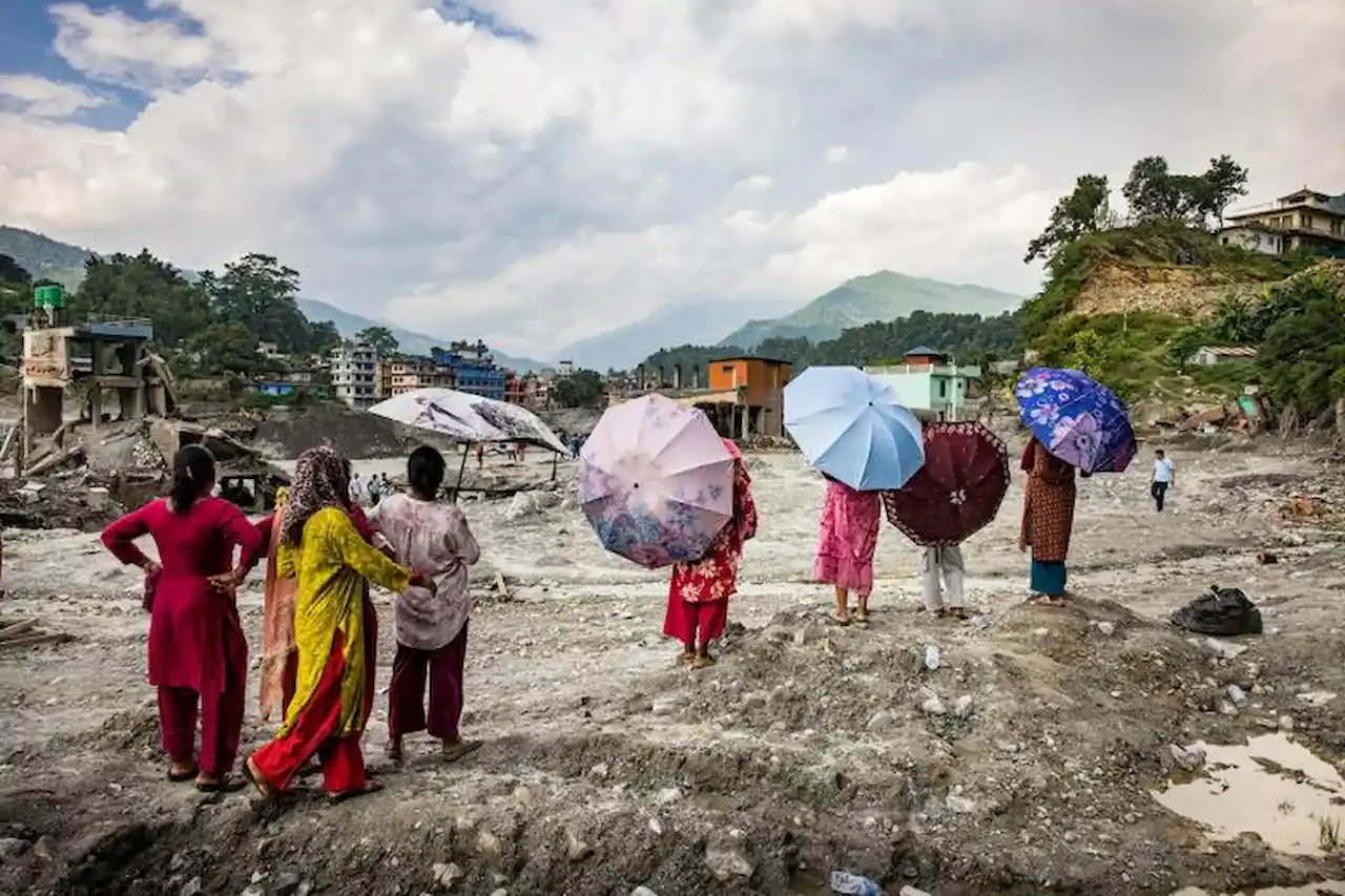 Varias personas con paraguas observan un terreno cubierto de escombros junto a una localidad de montaña / Foto: Jitendra Raj Bajracharya – ICIMOD